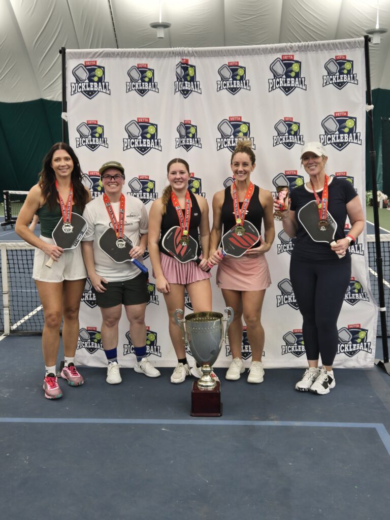 Five women stand side by side on a pickleball court, each holding paddles and wearing medals. A large trophy sits in front of them, celebrating their victory as 2025 Dink in the New Year Tourney Pickleball Champs. A tournament banner hangs behind.