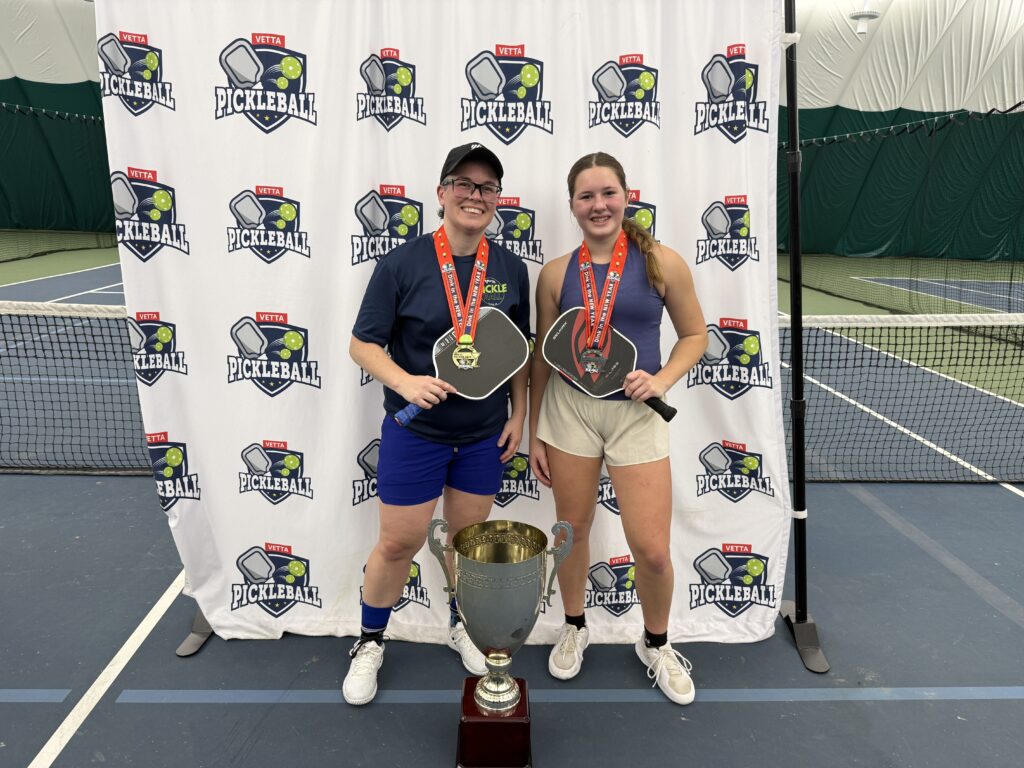 Two people stand on an indoor tennis court holding paddles, medals, and a large trophy. They are smiling in front of a “Pickleball” backdrop as the 2025 Dink in the New Year Tourney Pickleball Champs.