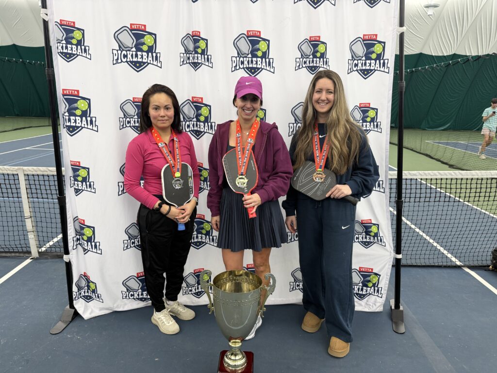 Three women stand on an indoor tennis court holding paddles and wearing medals in front of a 2025 Dink in the New Year Tourney Pickleball Champs banner. A large trophy is placed on the ground in front of them.