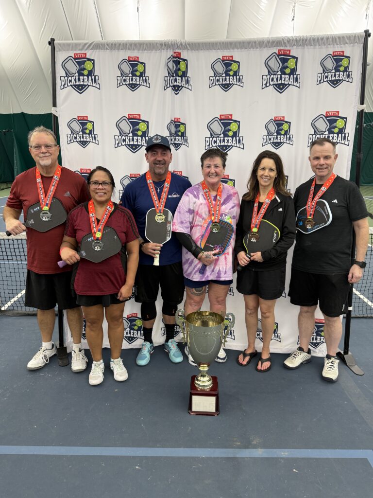 Six people wearing medals and holding pickleball paddles stand in front of a Pickleball banner on an indoor court. Smiling, the 2025 Dink in the New Year Tourney Pickleball Champs pose with their trophy, celebrating their victory.