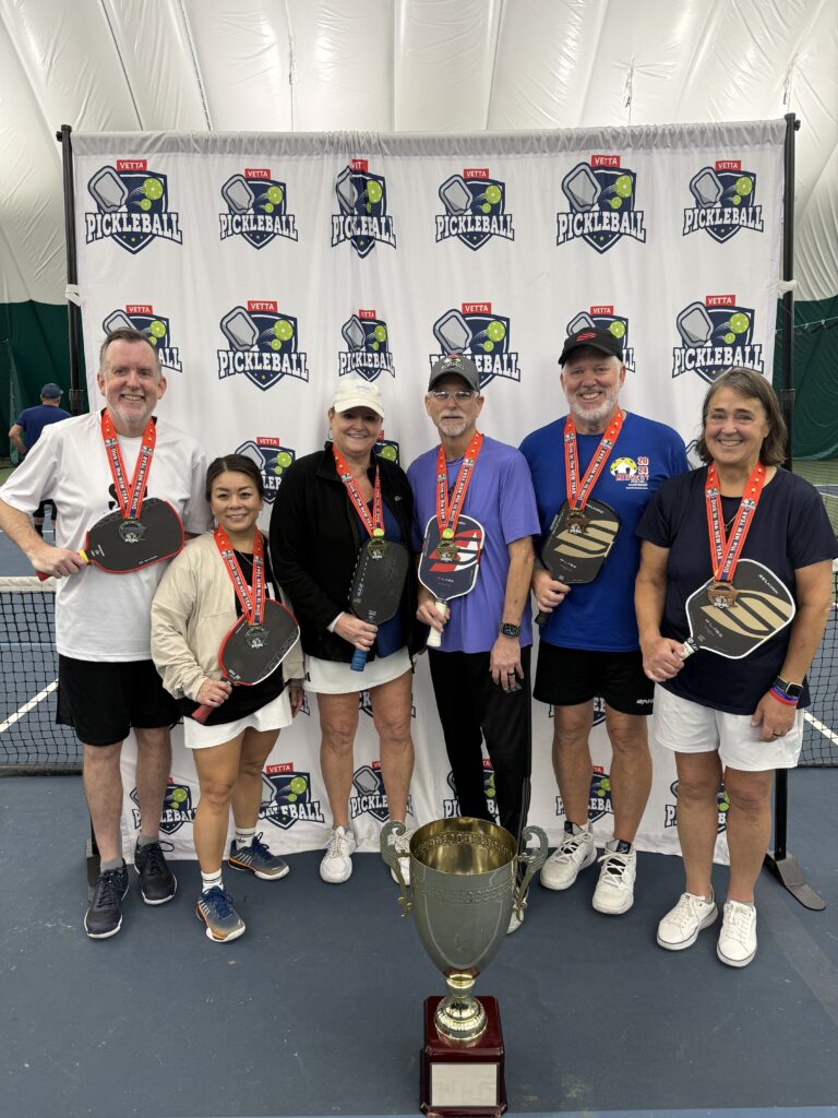 Six smiling 2025 Dink in the New Year Tourney Pickleball Champs, wearing medals, hold paddles and stand before a USA Pickleball banner on an indoor court. A large trophy sits proudly in front of the group.