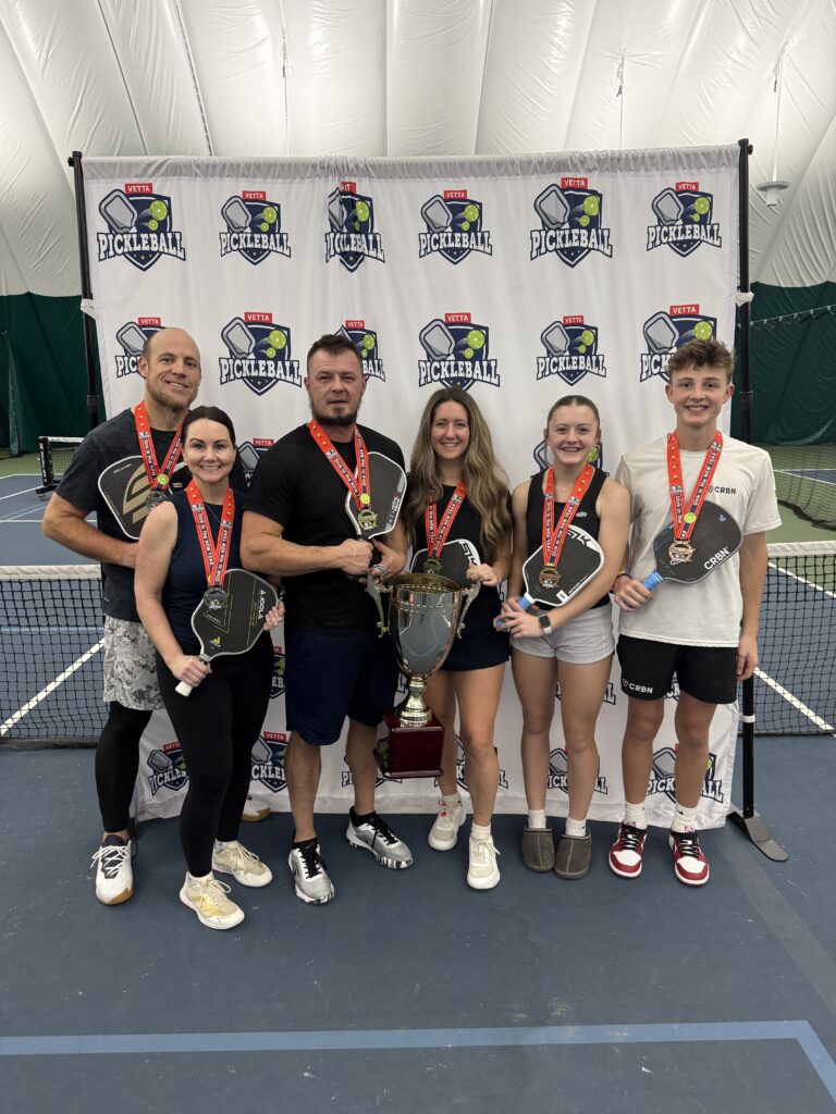 Six people with medals and paddles stand in front of a Pickleball banner on an indoor tennis court, smiling and holding a large trophy, celebrating their victory as 2025 Dink in the New Year Tourney Pickleball Champs.