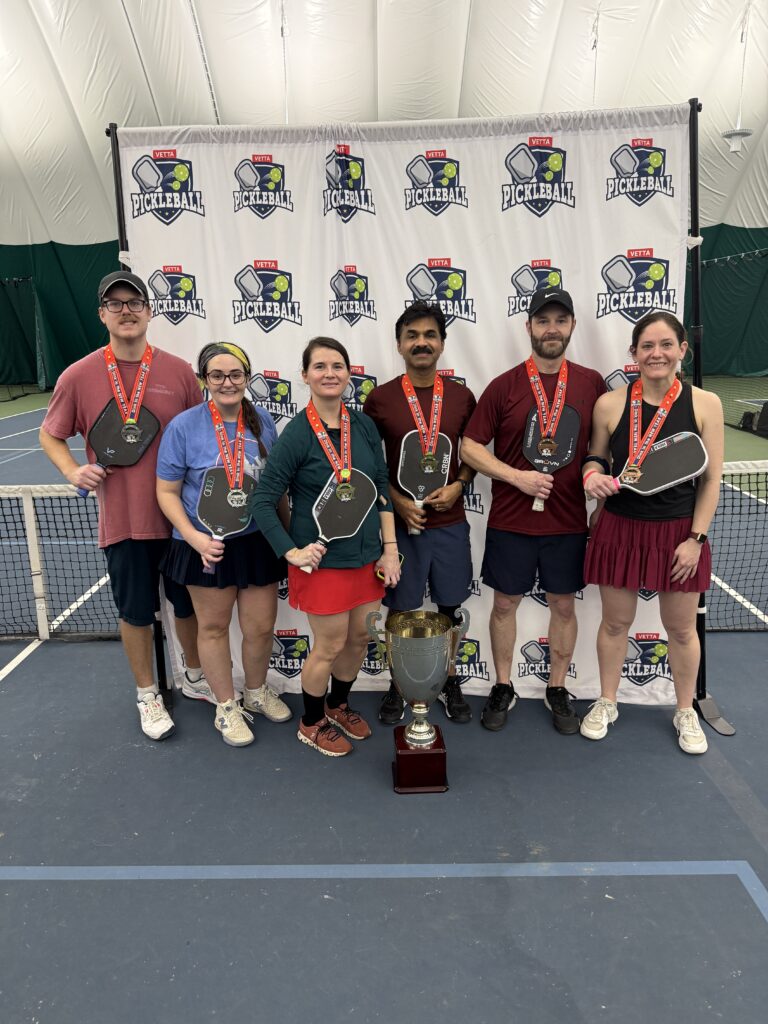 Six people stand indoors on a tennis court, smiling and holding pickleball paddles and medals. The 2025 Dink in the New Year Tourney Pickleball Champs pose proudly behind a large trophy and in front of a backdrop with pickleball logos.