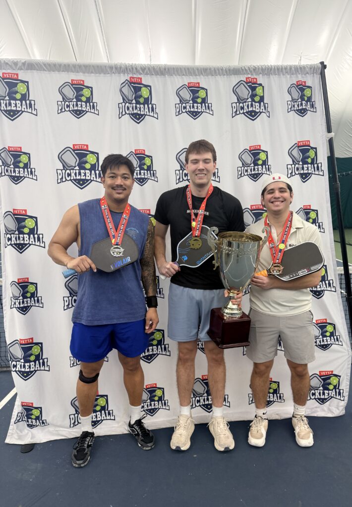 Three men stand in front of a Pickleball-branded backdrop, each wearing a medal and holding a paddle. The man in the center also holds a large trophy. All are smiling, celebrating as 2025 Dink in the New Year Tourney Pickleball Champs.