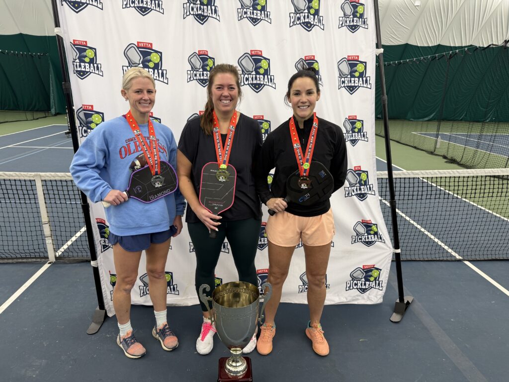 Three smiling women, the 2025 Dink in the New Year Tourney Pickleball Champs, stand on a tennis court with medals around their necks, holding paddles and posing proudly behind a large trophy and a Pickleball-branded backdrop.