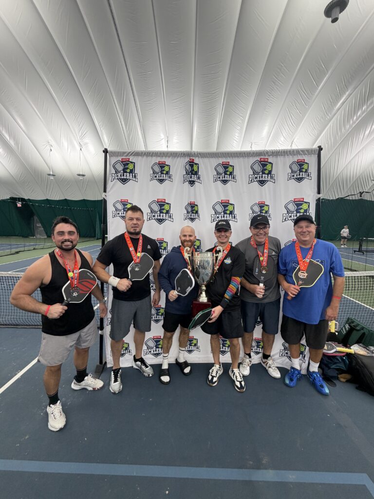 Six men stand on an indoor pickleball court, smiling and holding paddles. All wear medals, and one holds a large trophy. A Pickleball backdrop highlights these 2025 Dink in the New Year Tourney Pickleball Champs.