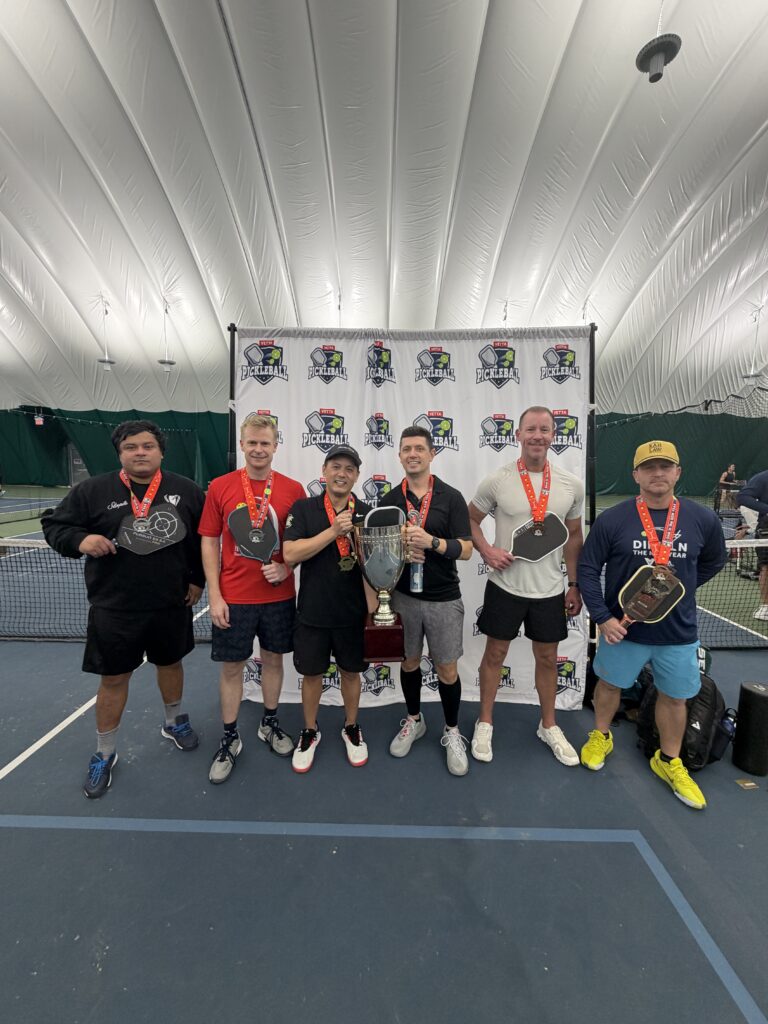 Six men wearing medals stand on an indoor tennis court, holding paddles and a trophy, posing in front of a Pickleball tournament backdrop. These 2025 Dink in the New Year Tourney Pickleball Champs are dressed in sportswear with shorts and sneakers.