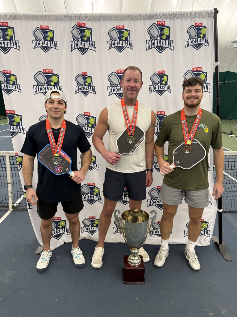 Three men wearing medals and holding pickleball paddles stand indoors in front of a Pickleball backdrop, smiling and posing with a large trophy at the 2025 Dink in the New Year Tourney Pickleball Champs event.