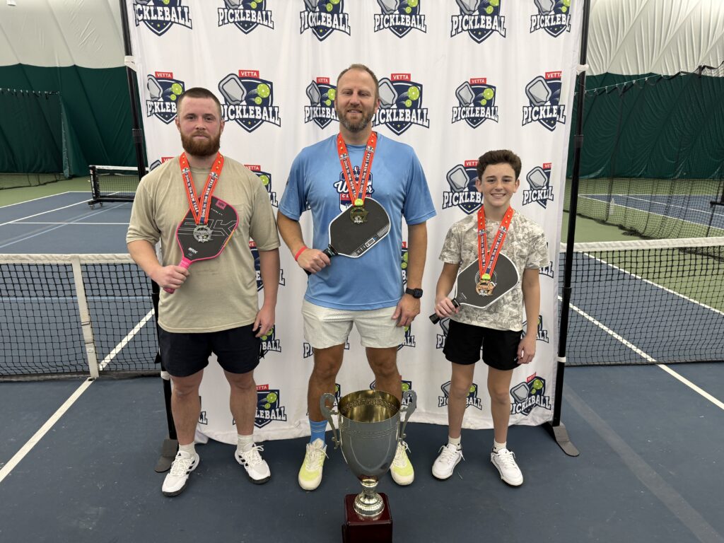 Three people stand on an indoor pickleball court, each holding a paddle and wearing a medal. Behind them is a Pickleball-branded backdrop. A large trophy sits in front, celebrating the 2025 Dink in the New Year Tourney Pickleball Champs.