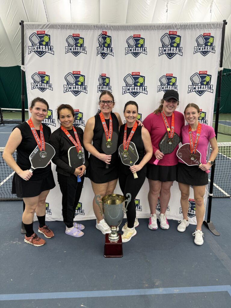 Six women wearing medals and holding pickleball paddles stand in front of a "2025 Dink in the New Year Tourney" banner, smiling proudly. A large trophy sits before these Pickleball Champs on the indoor court.