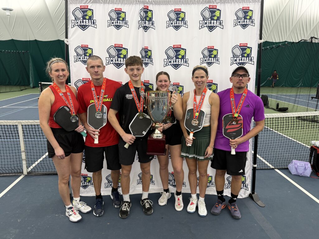 Six pickleball players stand on an indoor court, smiling and holding medals. Centered around a trophy with a Pickleball banner behind them, they proudly celebrate as the 2025 Dink in the New Year Tourney Pickleball Champs.