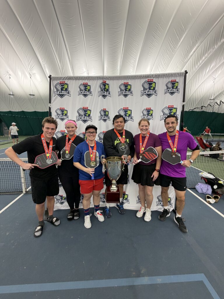 Six people stand on an indoor pickleball court, smiling and wearing medals around their necks. As 2025 Dink in the New Year Tourney Pickleball Champs, they hold paddles and a large trophy, posing in front of a banner with pickleball logos.