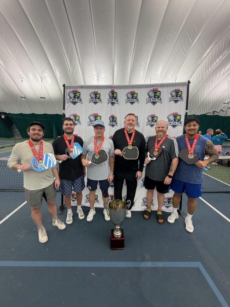 Six men stand on an indoor pickleball court in front of a Pickleball banner, wearing medals and smiling. Some hold paddles, with a large trophy at their feet—true 2025 Dink in the New Year Tourney Pickleball Champs.