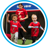 Two young children in red shirts run and smile on a field, while an adult in a matching shirt follows behind them, also smiling. All appear to be having fun, participating in an indoor sports activity.
