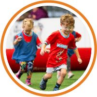 Two young children run on a grassy field during a sports activity, both wearing colorful outfits and focused expressions. A red foam barrier and blurred background are visible behind them.