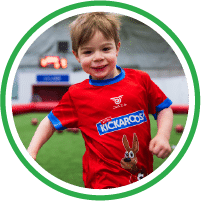 A young boy wearing a red T-shirt with a cartoon rabbit is smiling and running indoors on an artificial turf field. Blurred soccer balls and a scoreboard are visible in the background.