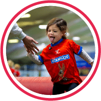 A young child in a red sports shirt reaches out for a high-five from an adult, smiling and playing indoors.