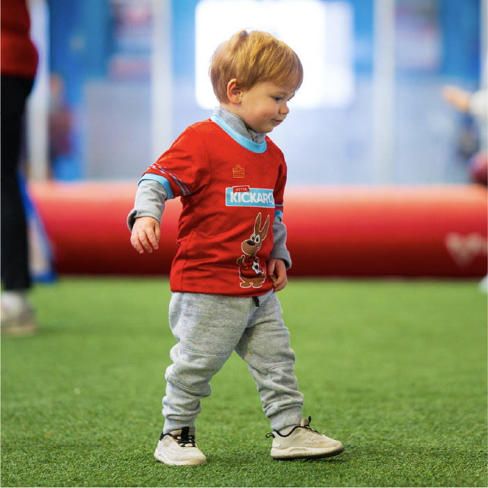Toddler in a red Kickaroo shirt and gray pants walks on indoor turf, with blurred people and a red object in the background.