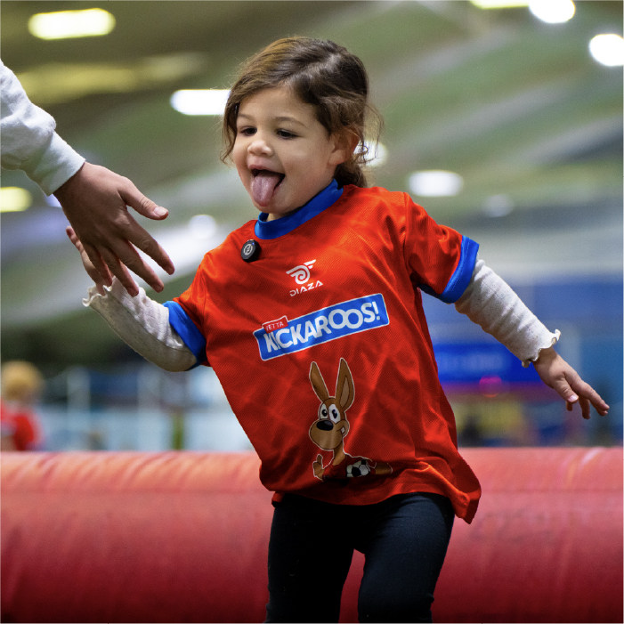A young child in a red Kickaroos! shirt sticks out her tongue and reaches for an adults hand while playing indoors.