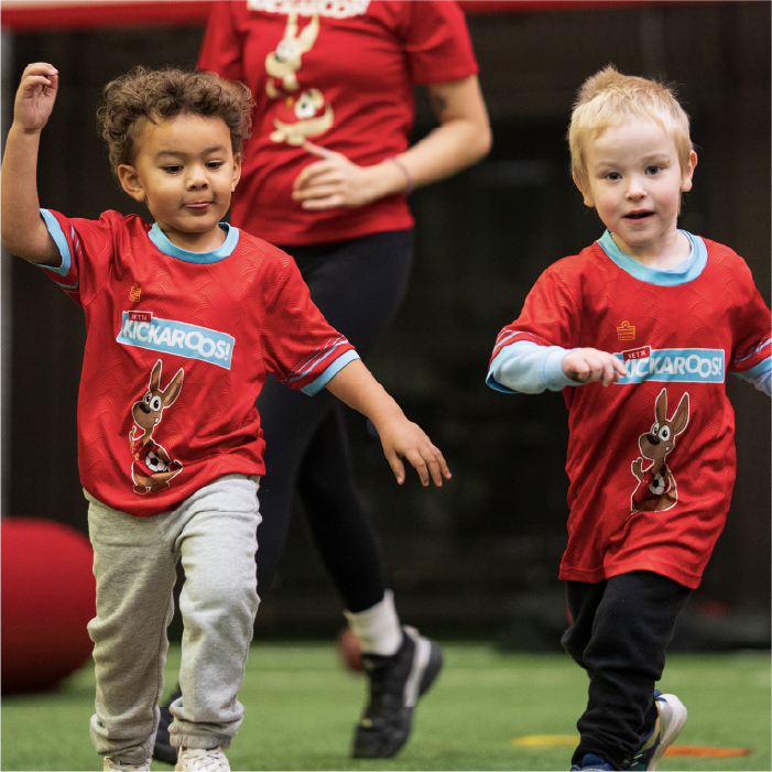 Two young children in red shirts run playfully indoors on green turf, smiling with arms raised. An adult stands behind them, partially visible. A large red ball sits on the turf nearby.