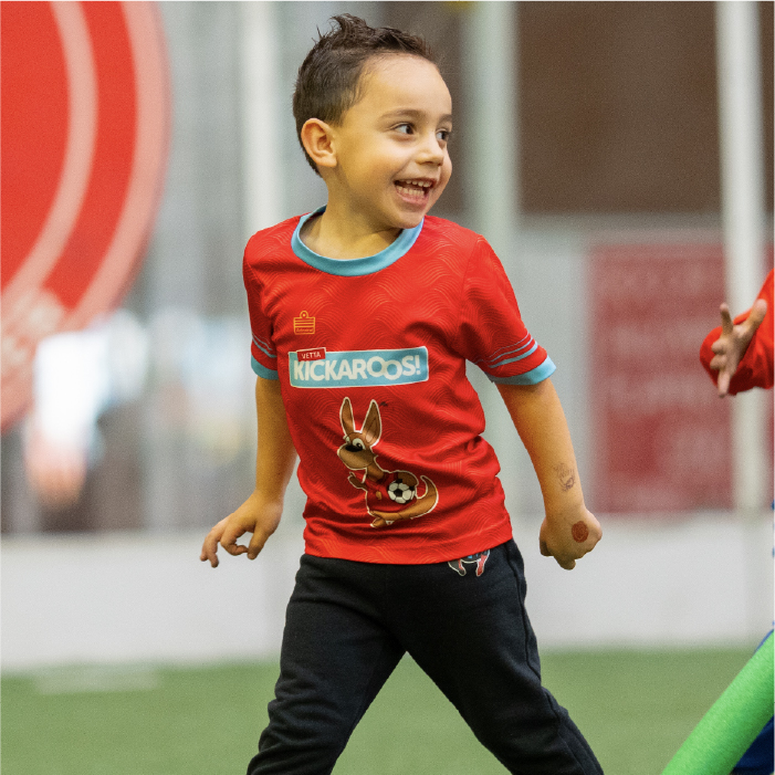 Smiling young child wearing a red Kickaroos shirt and black pants runs on indoor turf, looking to the side with joy.