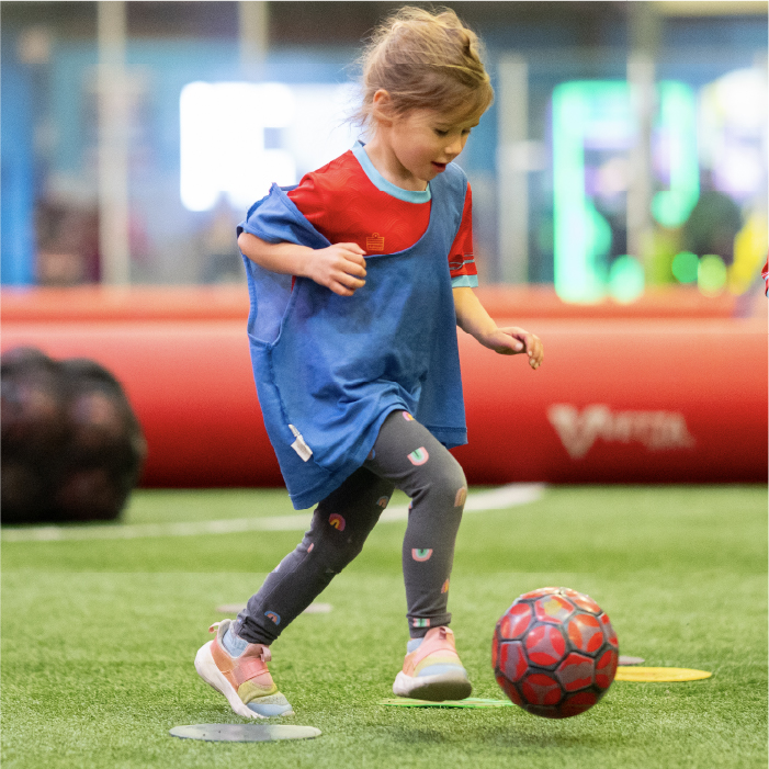 A young child wearing a blue and red sports jersey kicks a red soccer ball on indoor turf, focused on playing.