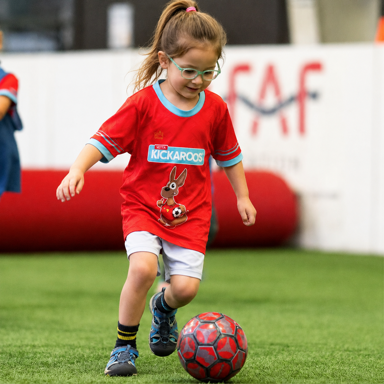 A young boy wearing glasses and a red shirt with a bunny design chases a red soccer ball on an indoor turf field.