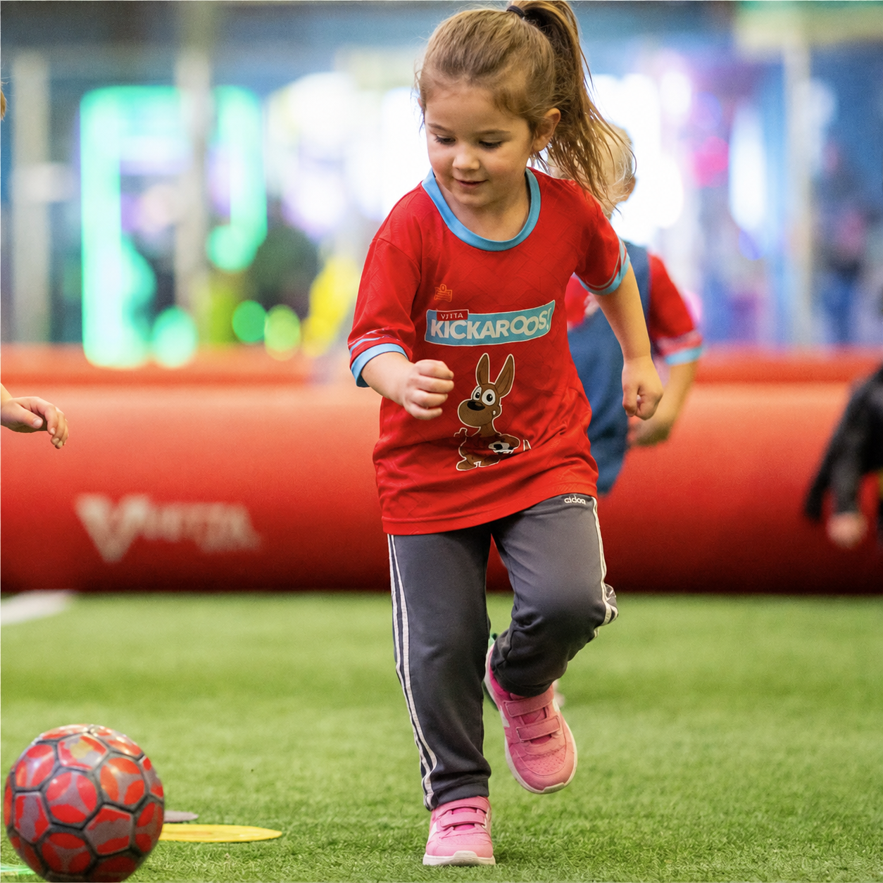 A young boy in a red shirt runs on indoor turf, smiling and chasing a red soccer ball, with other children and a red barrier visible in the background.