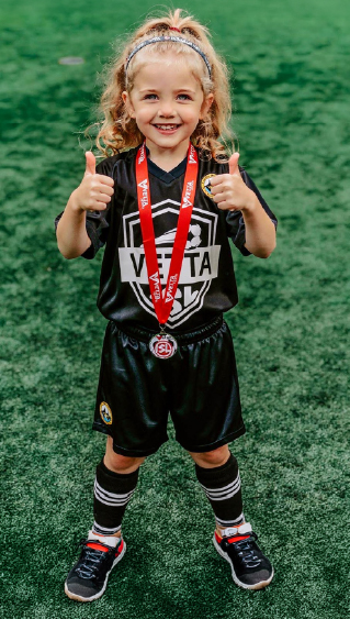 Smiling young child in a black soccer uniform stands on green turf, giving two thumbs up and wearing a red medal around their neck—a proud moment made possible by fun soccer lessons for toddlers.