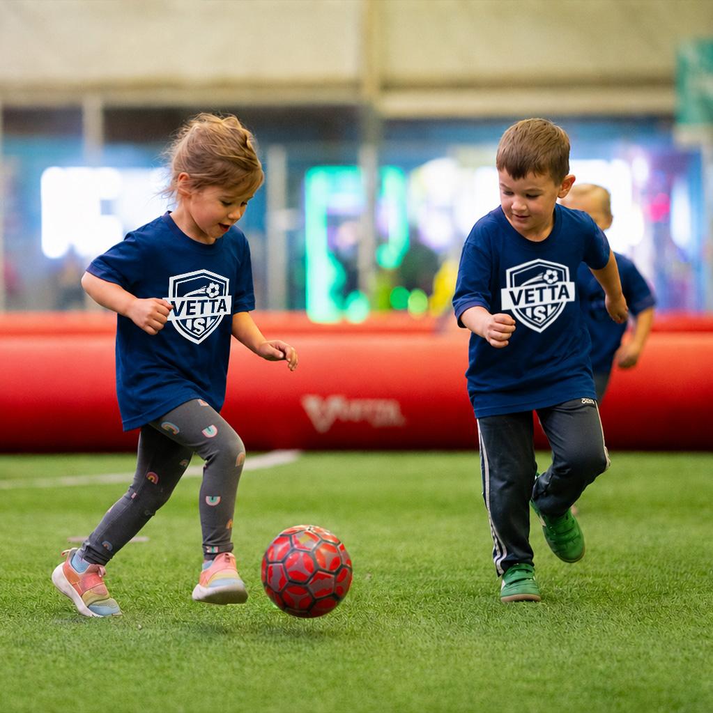 Toddler in a red Kickaroo shirt and gray pants walks on indoor turf, with blurred people and a red object in the background.
