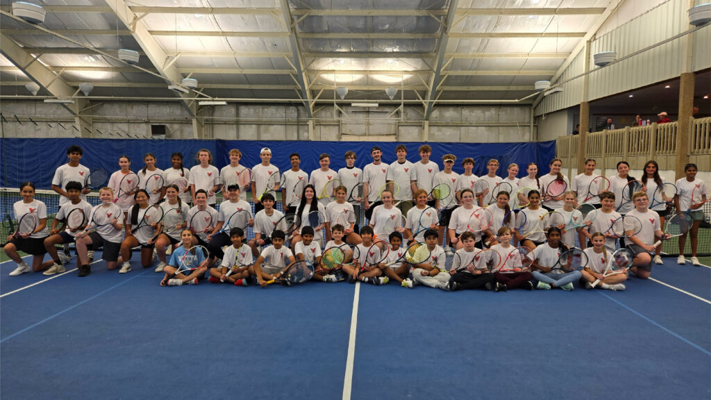 A large group of Vetta junior tennis players in white shirts posing on an indoor blue court holding their rackets.